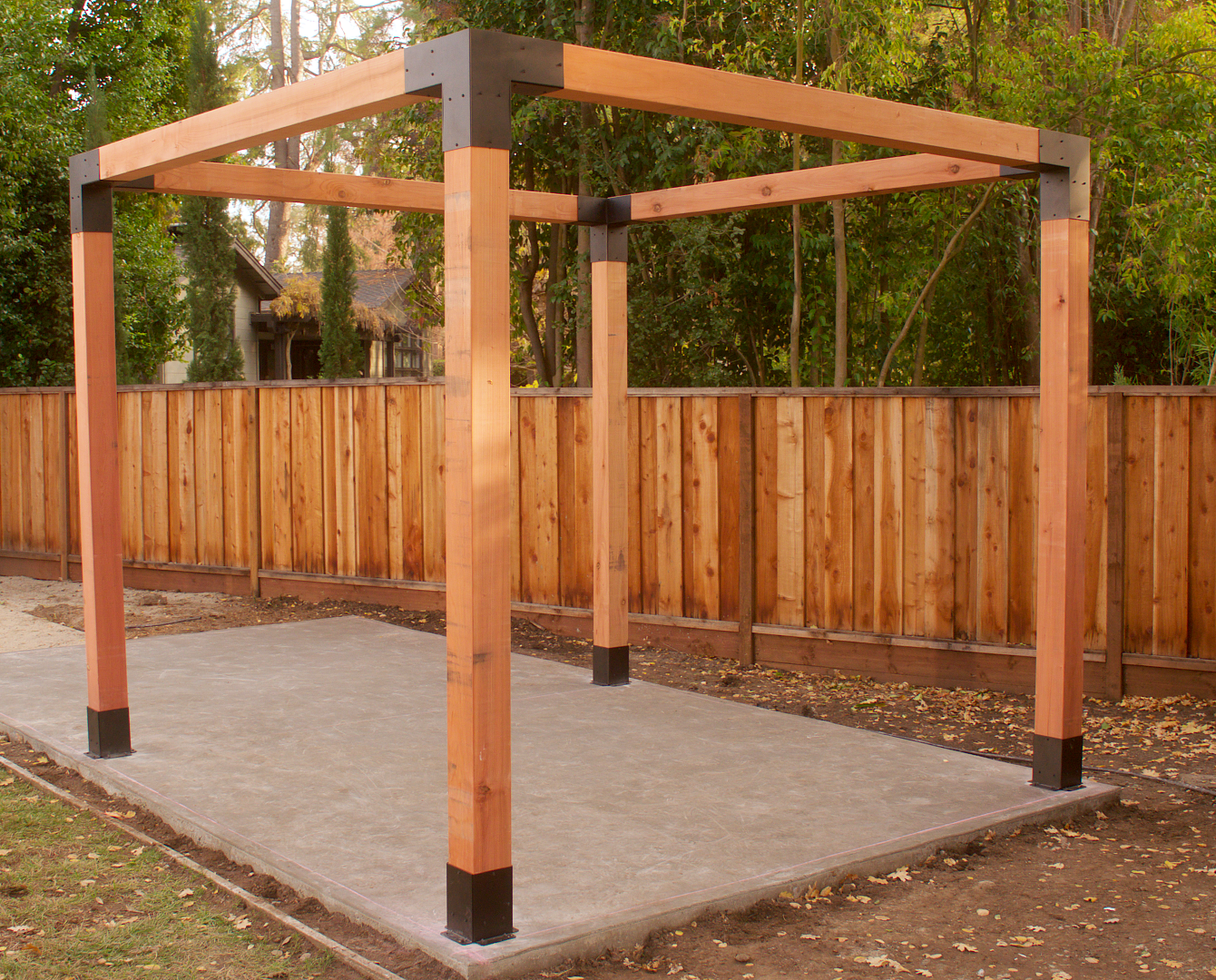 Completed Simpson Sage DIY Pergola structure on a concrete slab with a wooden fence and trees in the background