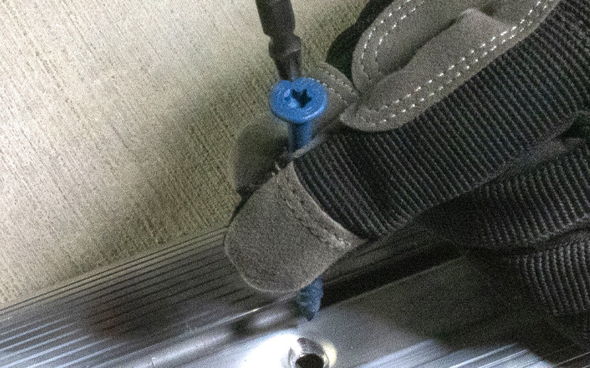 Hand wearing a glove holding a blue concrete screw near a metal surface
