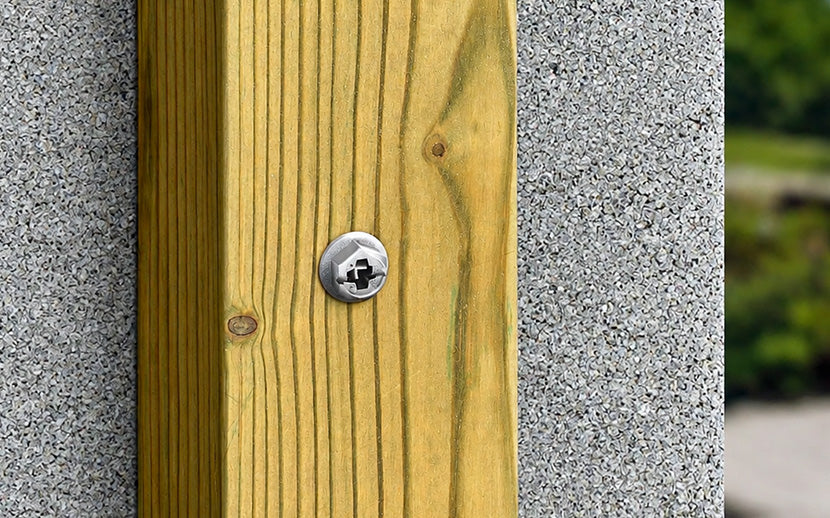 A close-up of a stainless steel concrete screw fastened to a vertical wooden post, set against a concrete wall