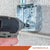 Person using a drill to fasten a metal box onto a concrete wall with 'Fasteners Plus' branding.