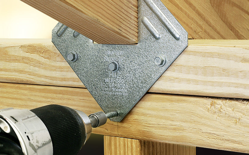 Metal corner brace on wooden beams with a drill bit inserted, indicating construction or woodworking activity.